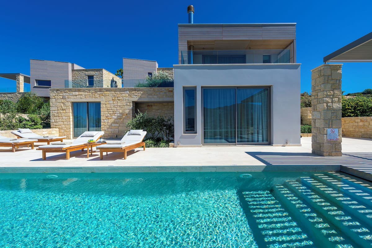 A modern poolside area with lounge chairs, a clear pool, and a contemporary building with large windows and stone walls.