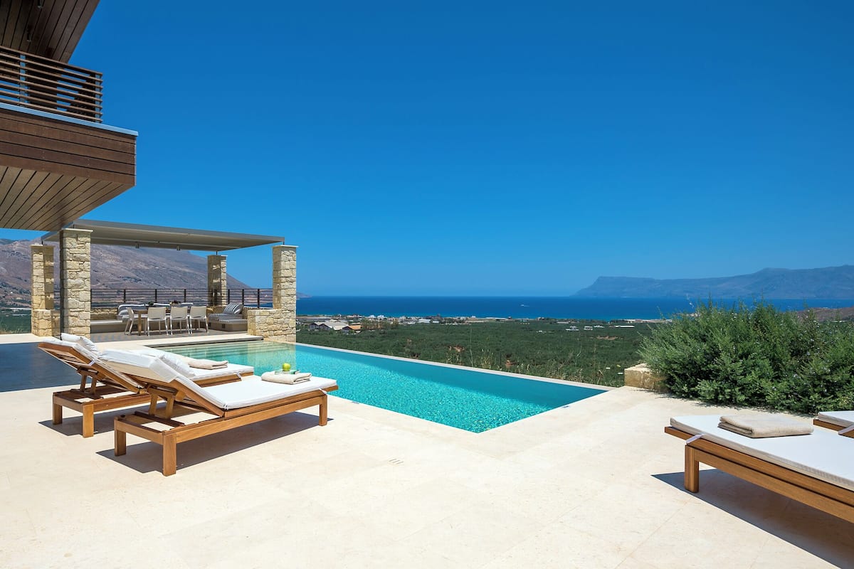 A poolside area with lounge chairs, a stone structure, and a view of the sea and mountains.