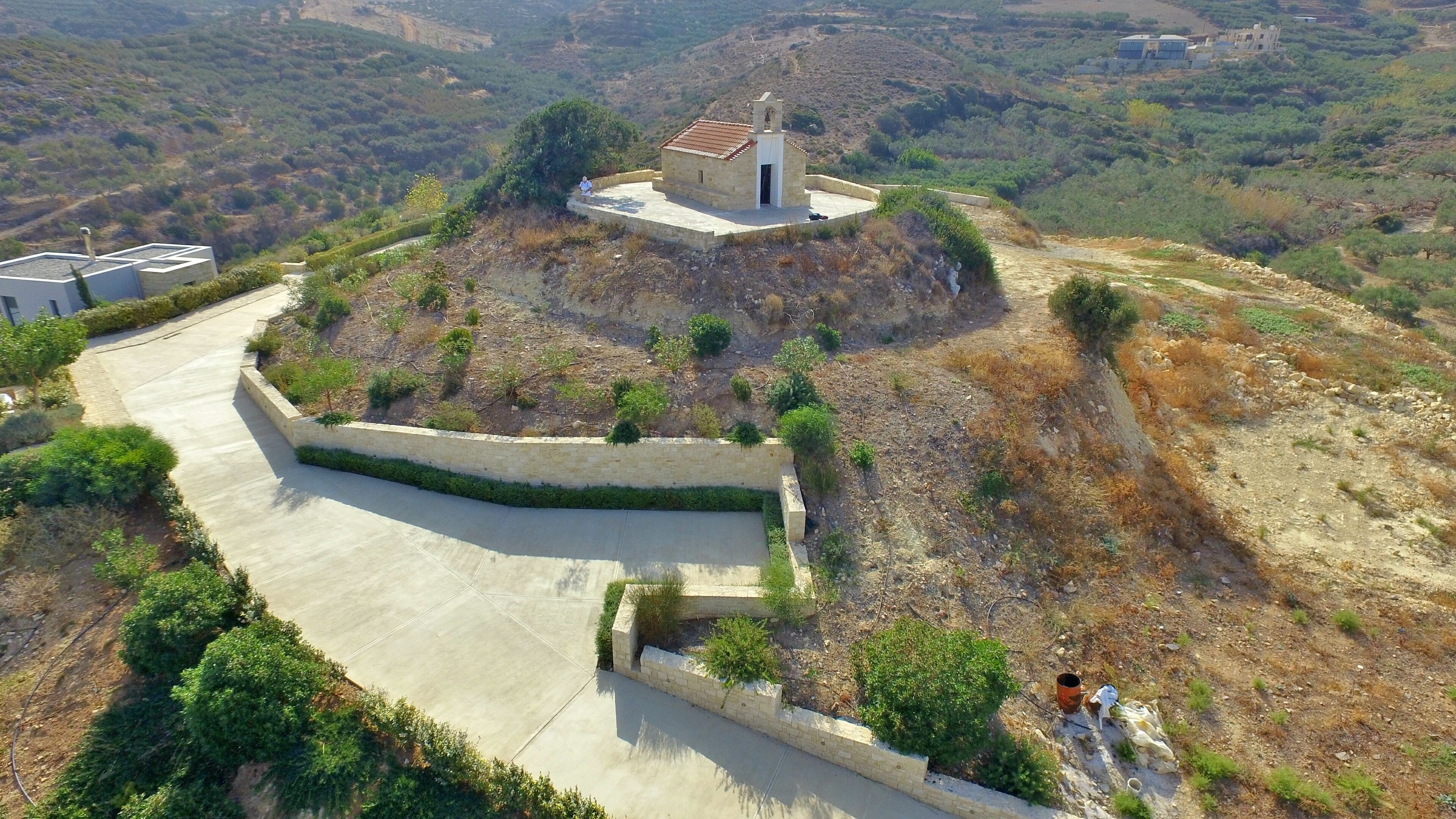 A small stone chapel with a bell tower, surrounded by a landscaped area with a curved pathway and a retaining wall.
