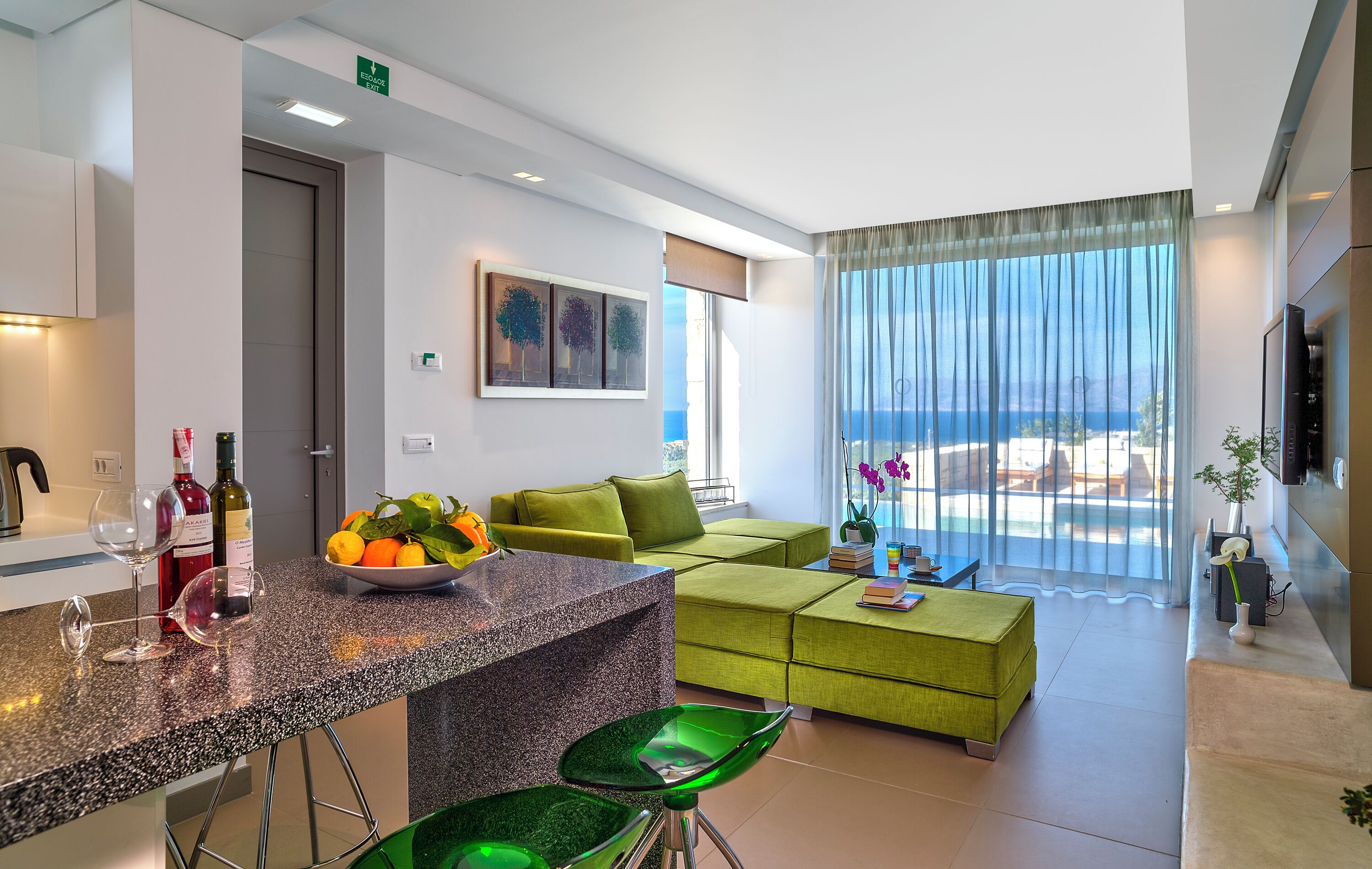A modern living room with a green sofa, a kitchen area with a granite countertop, and a view of the ocean through large windows.