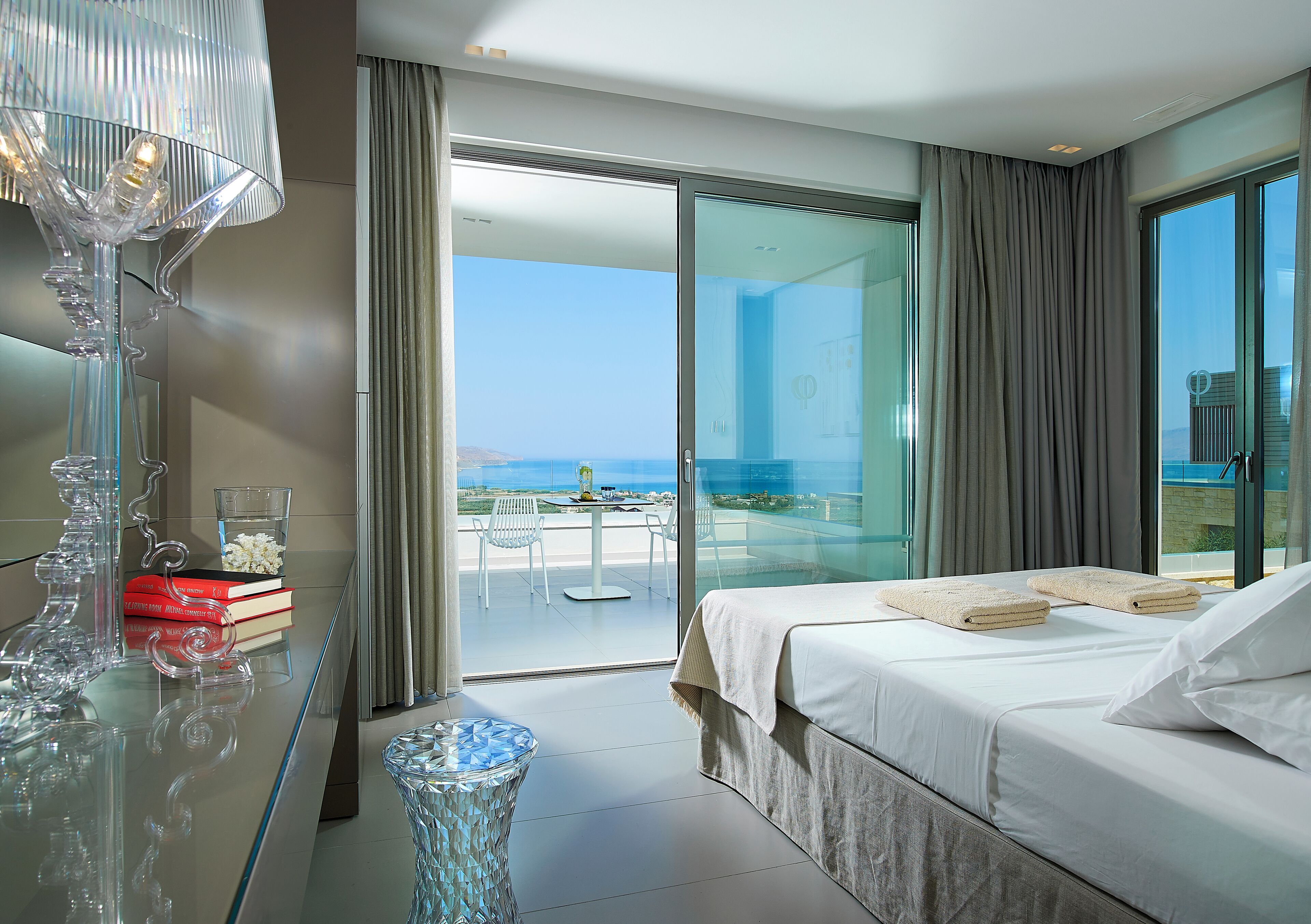 A modern hotel room with a large bed, a glass table with books, a decorative lamp, and a view of the sea through large windows.