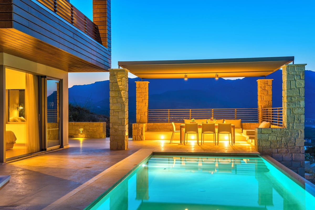 A modern outdoor pool area with stone pillars, a covered seating area, and a view of mountains.
