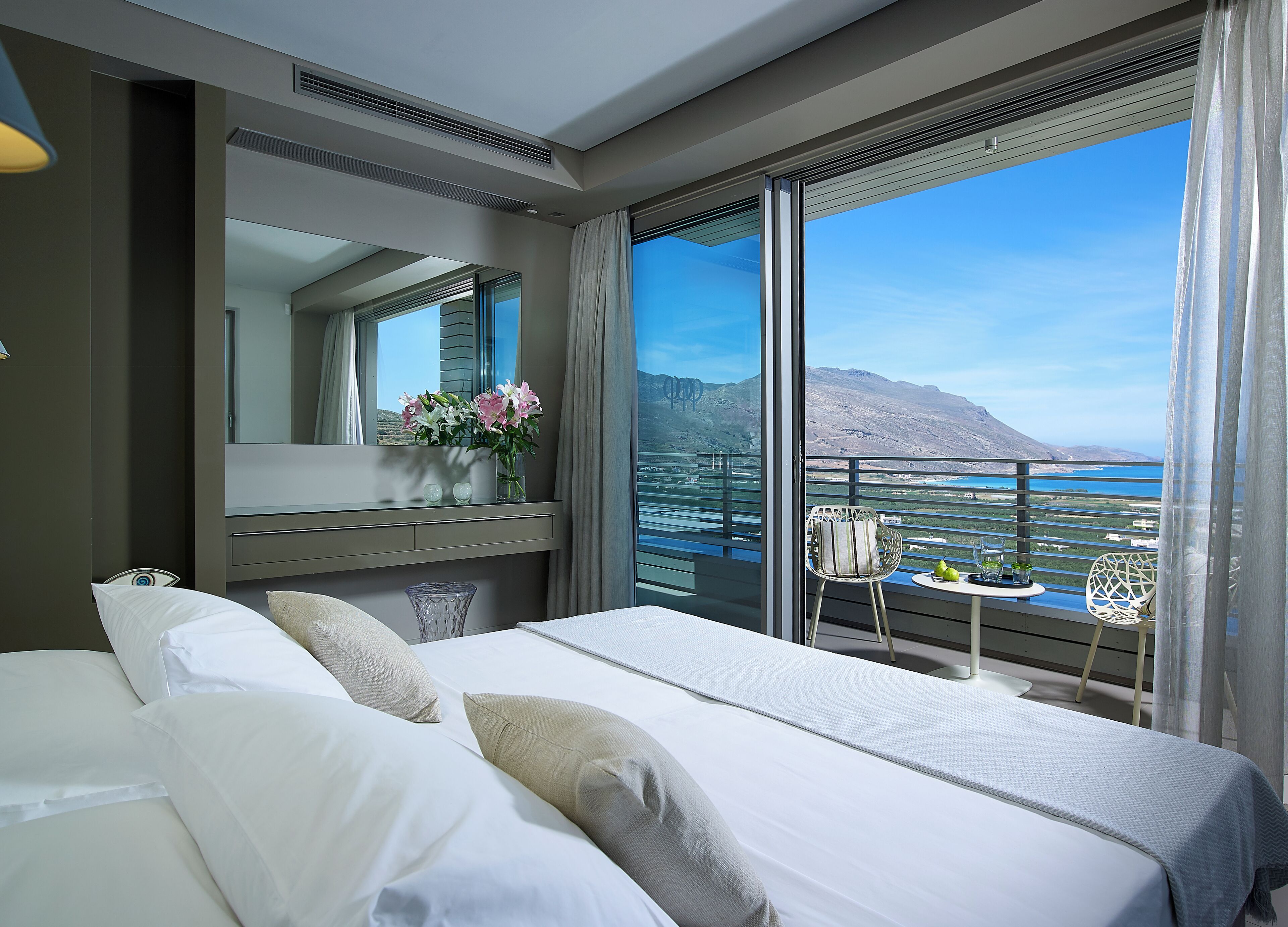 A hotel room with a large bed, a vanity with a mirror, a small table with a plant, and a balcony with a view of the sea and mountains.
