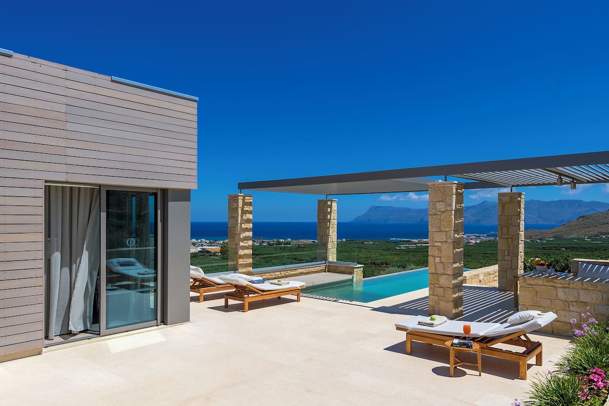 A modern outdoor pool area with lounge chairs, a table with a drink, and a view of the sea and mountains.