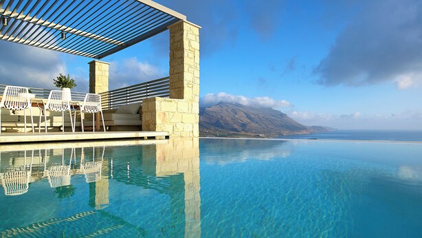 An infinity pool with clear blue water, a stone structure, and white chairs under a pergola.