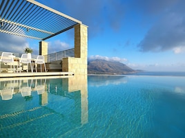 An infinity pool with clear blue water, a stone structure, and white chairs under a pergola.