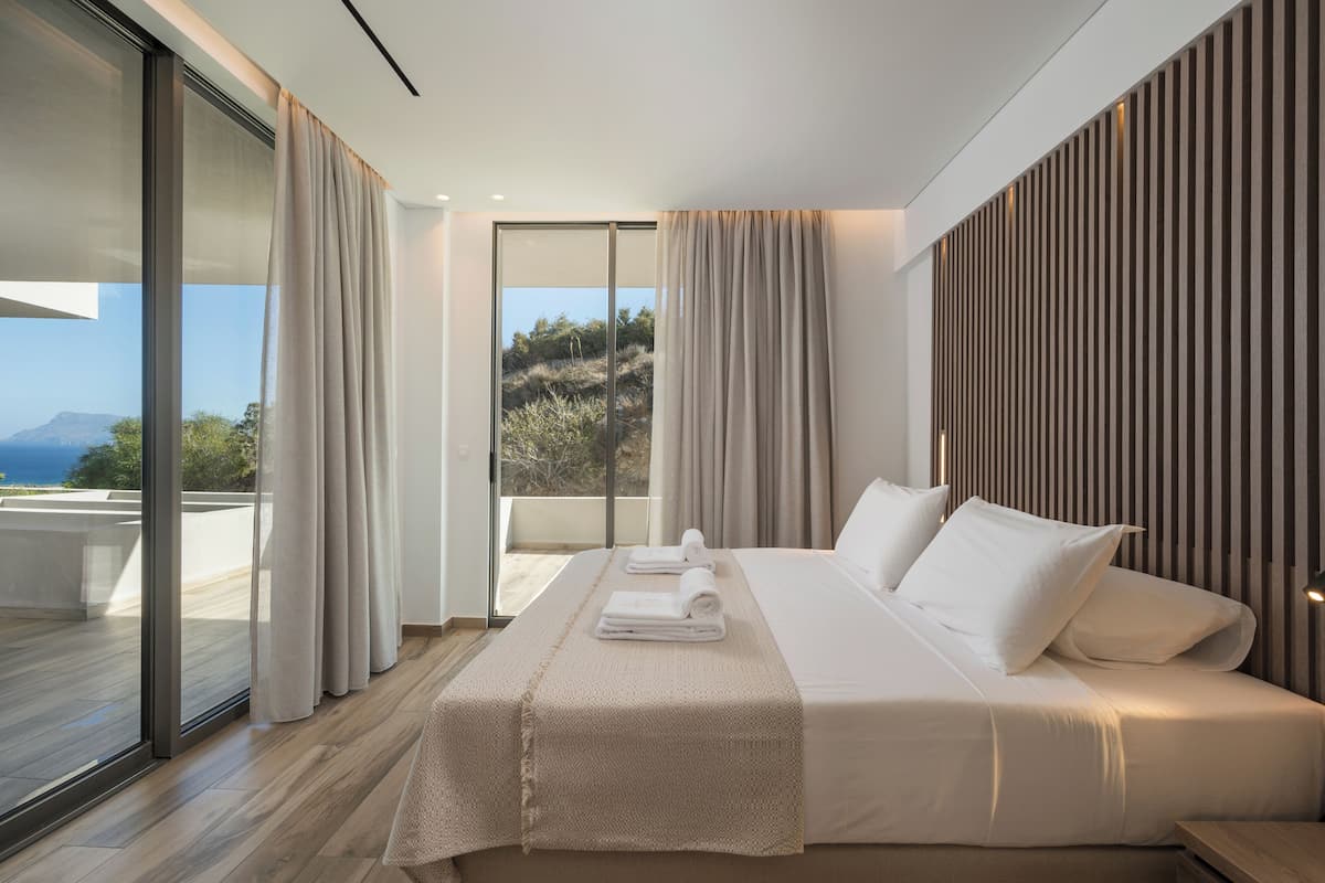 A modern hotel room with a large bed, a balcony with a view of the sea, and a sliding glass door.