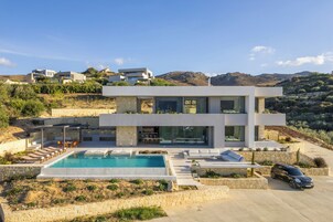Una casa moderna de dos pisos con piscina, mobiliario exterior y un coche estacionado frente a ella.