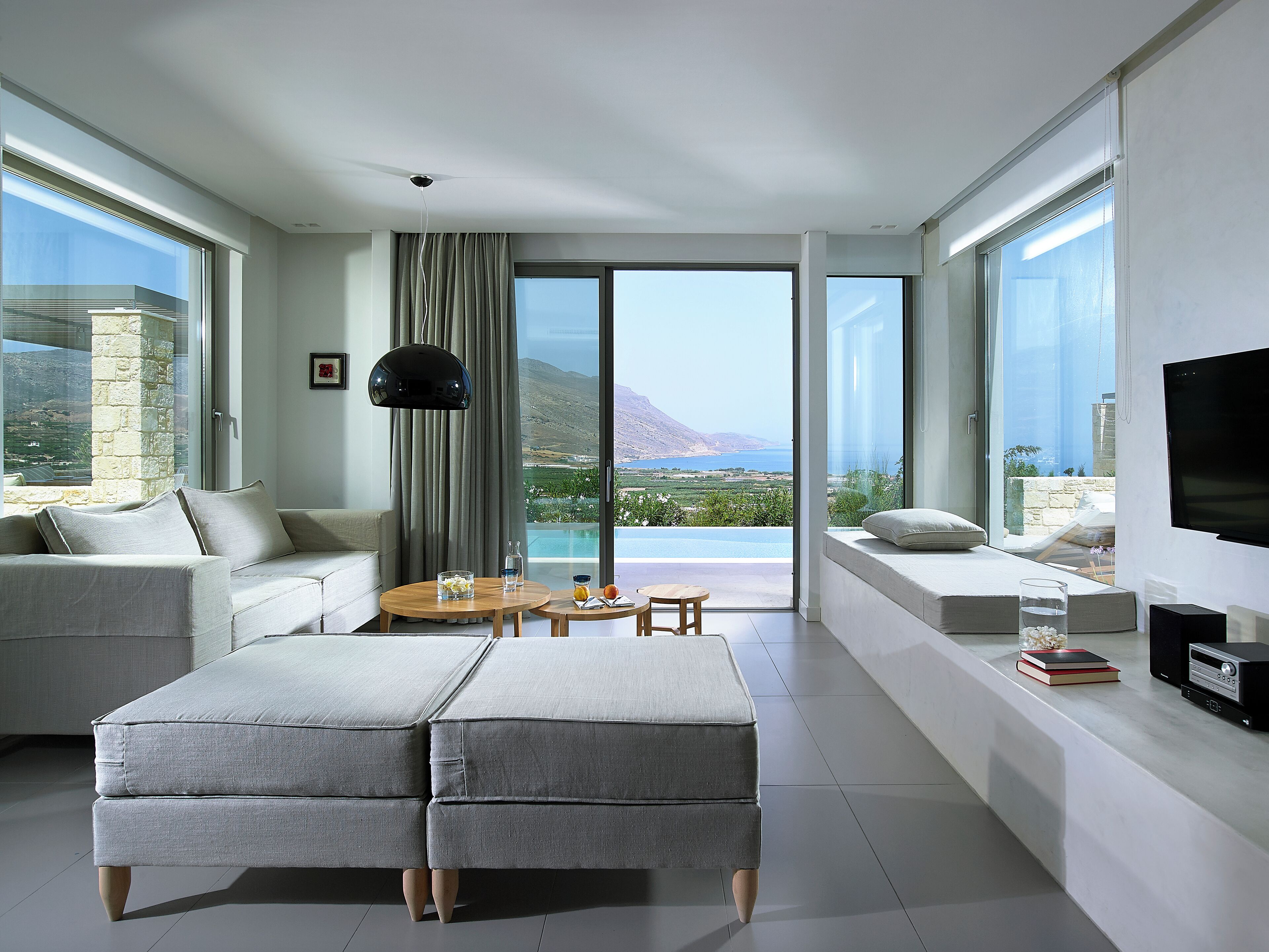 A modern living room with a sofa, ottoman, and a dining table with chairs. There is a large window with a view of a mountainous landscape and a pool.