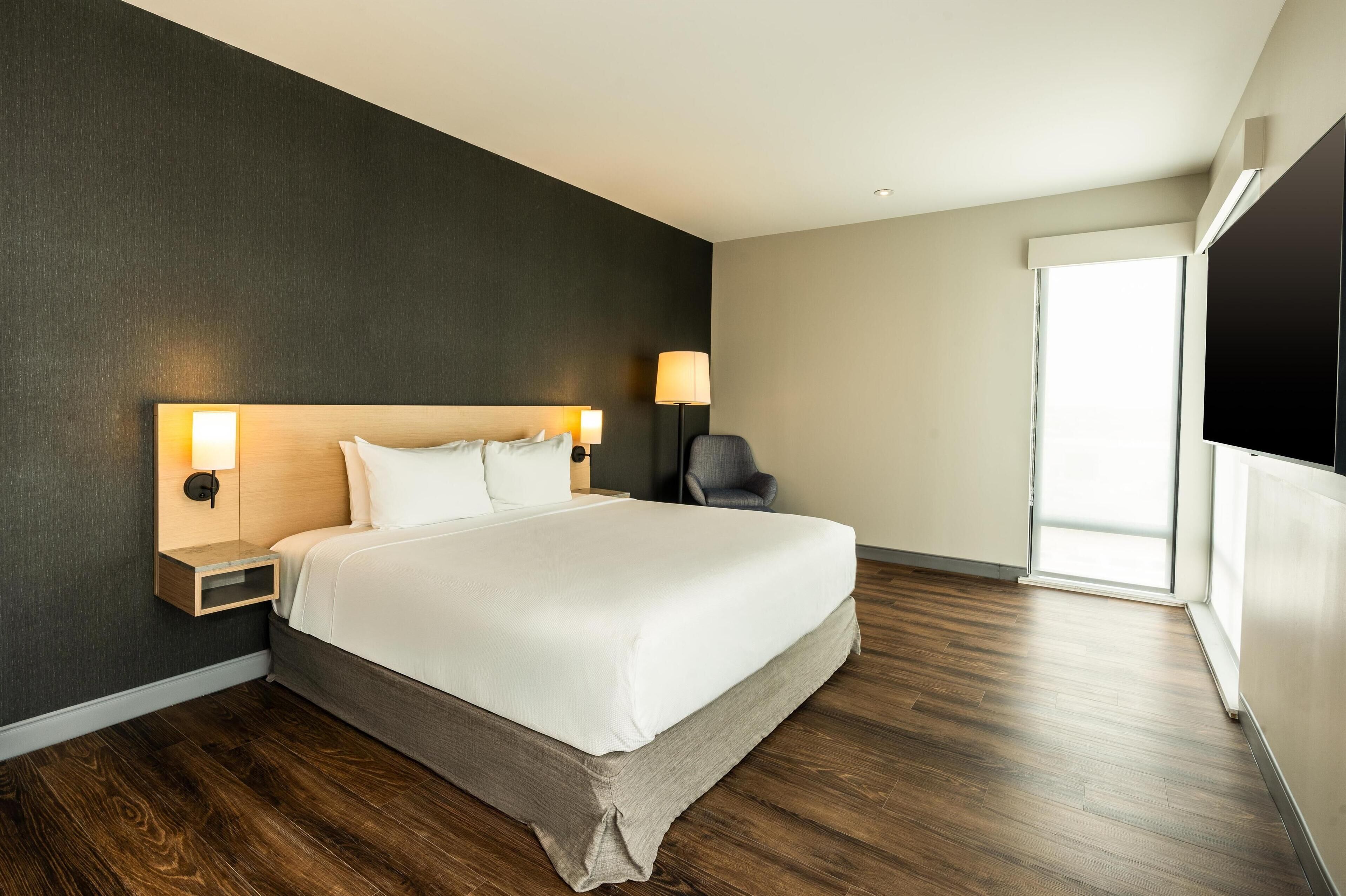 A modern hotel room with a large bed, dark walls, and a large window.