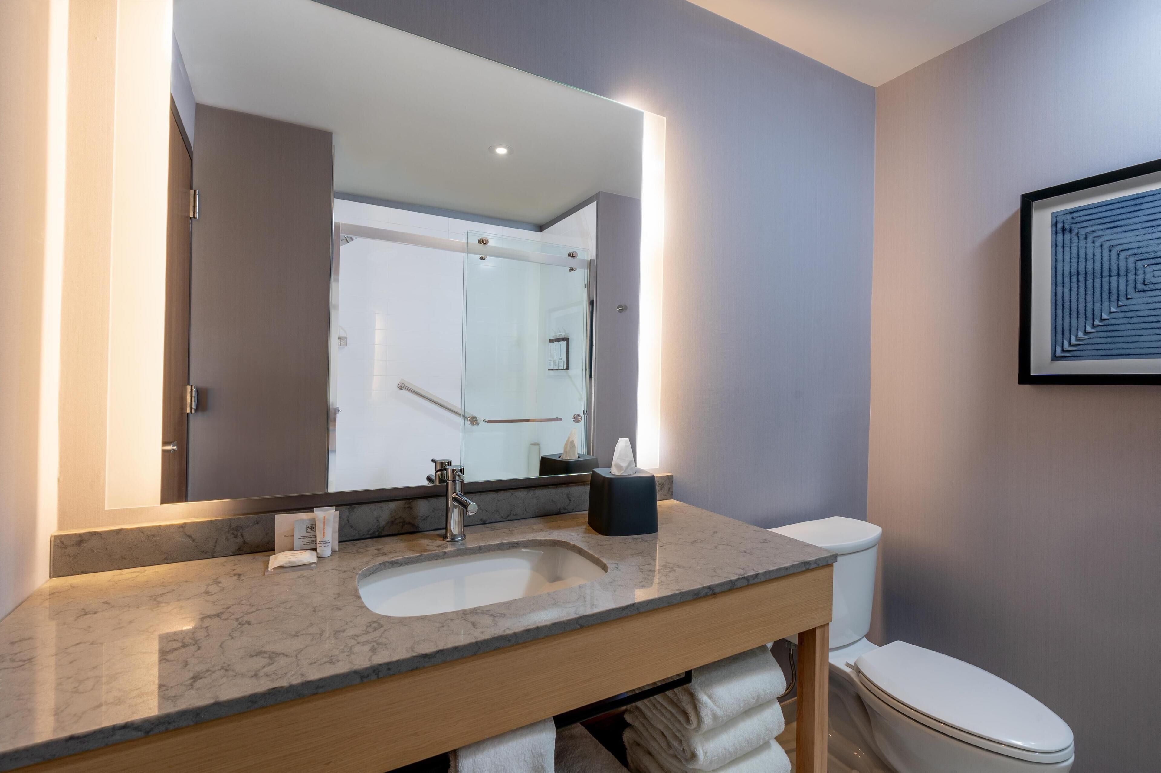 A modern bathroom with a marble sink, a large mirror, a framed artwork, and a visible shower area.