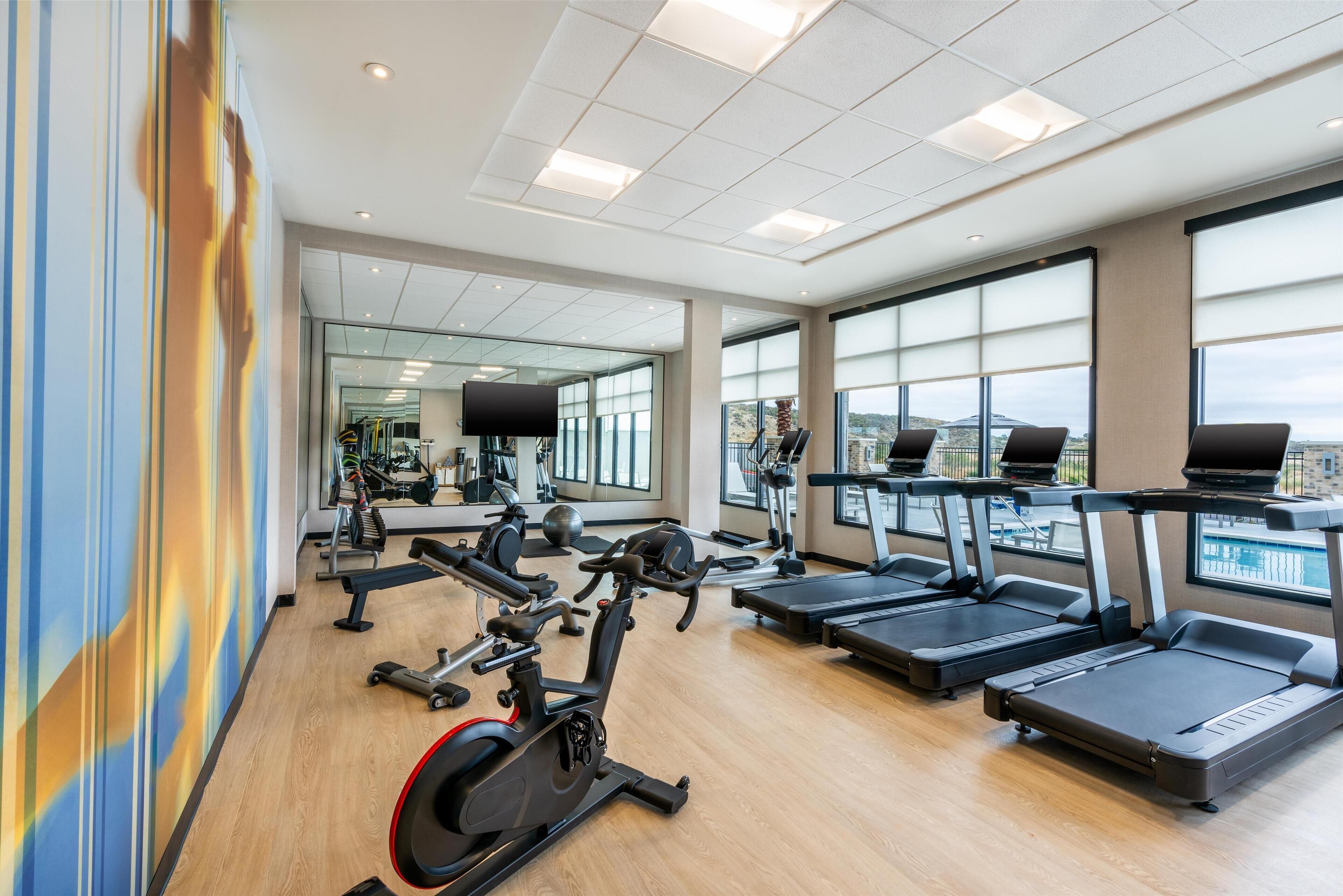 Une salle de sport équipée de nombreux appareils d’exercice, dotée de grandes fenêtres et offrant une vue sur la ville.