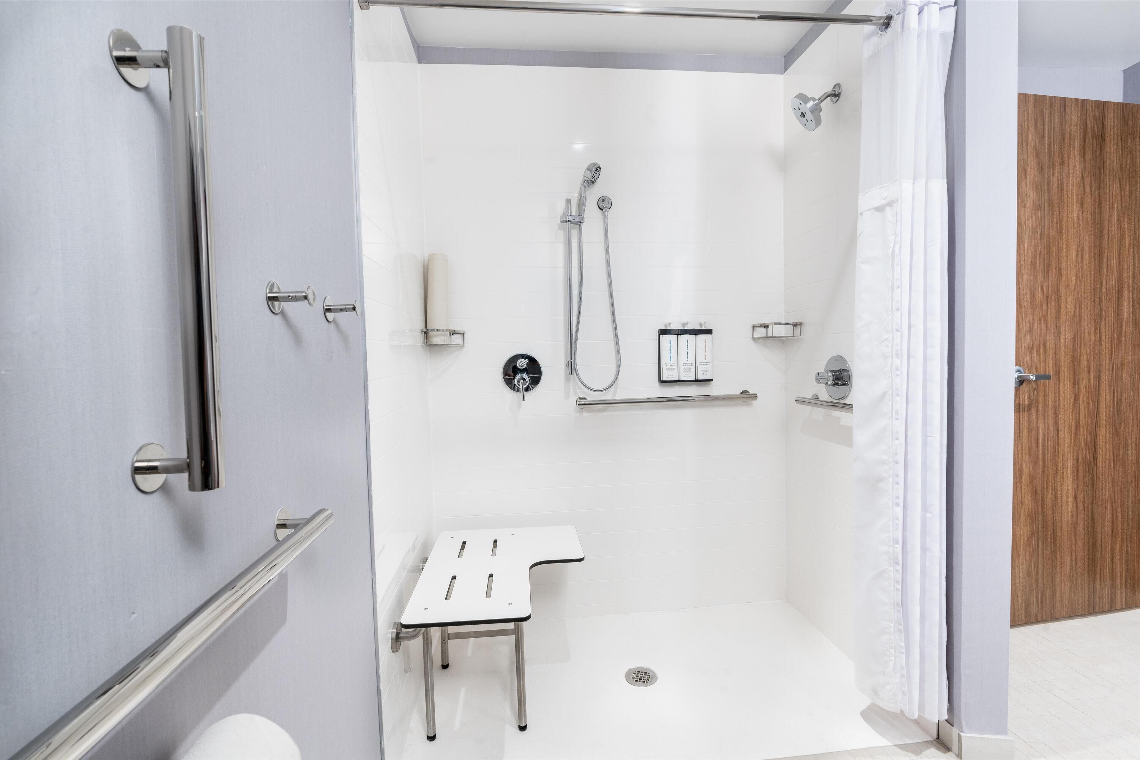 Un área de ducha moderna con puerta de vidrio, un cabezal de ducha manual, un grifo manual, una mesita blanca pequeña y un perchero para toallas.