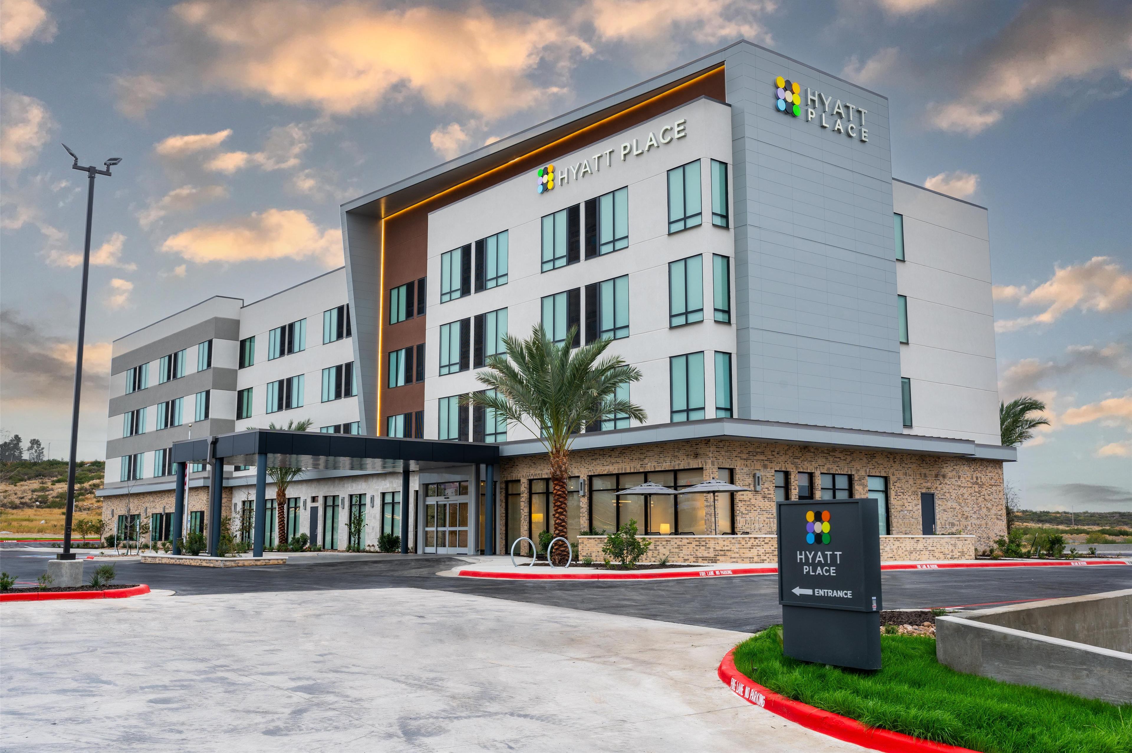 A modern Hyatt Place hotel with a prominent entrance, a landscaped area with palm trees, and a clear sky.