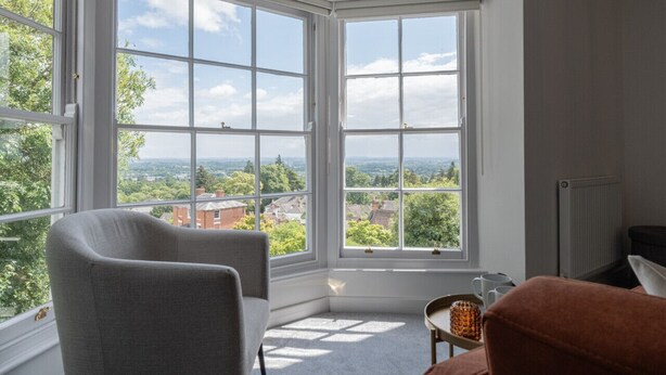 A room with large windows offering a view of a green landscape and buildings. There is a grey armchair and a brown sofa. - Cosy 1BD in Malvern With Stunning Views (Malvern)