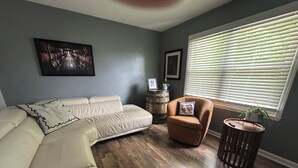 Un salon comprenant un canapé, un fauteuil, un tonneau en bois et une fenêtre avec des stores.