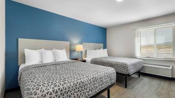 A hotel room with two beds, a nightstand with a lamp, and a window with blinds.