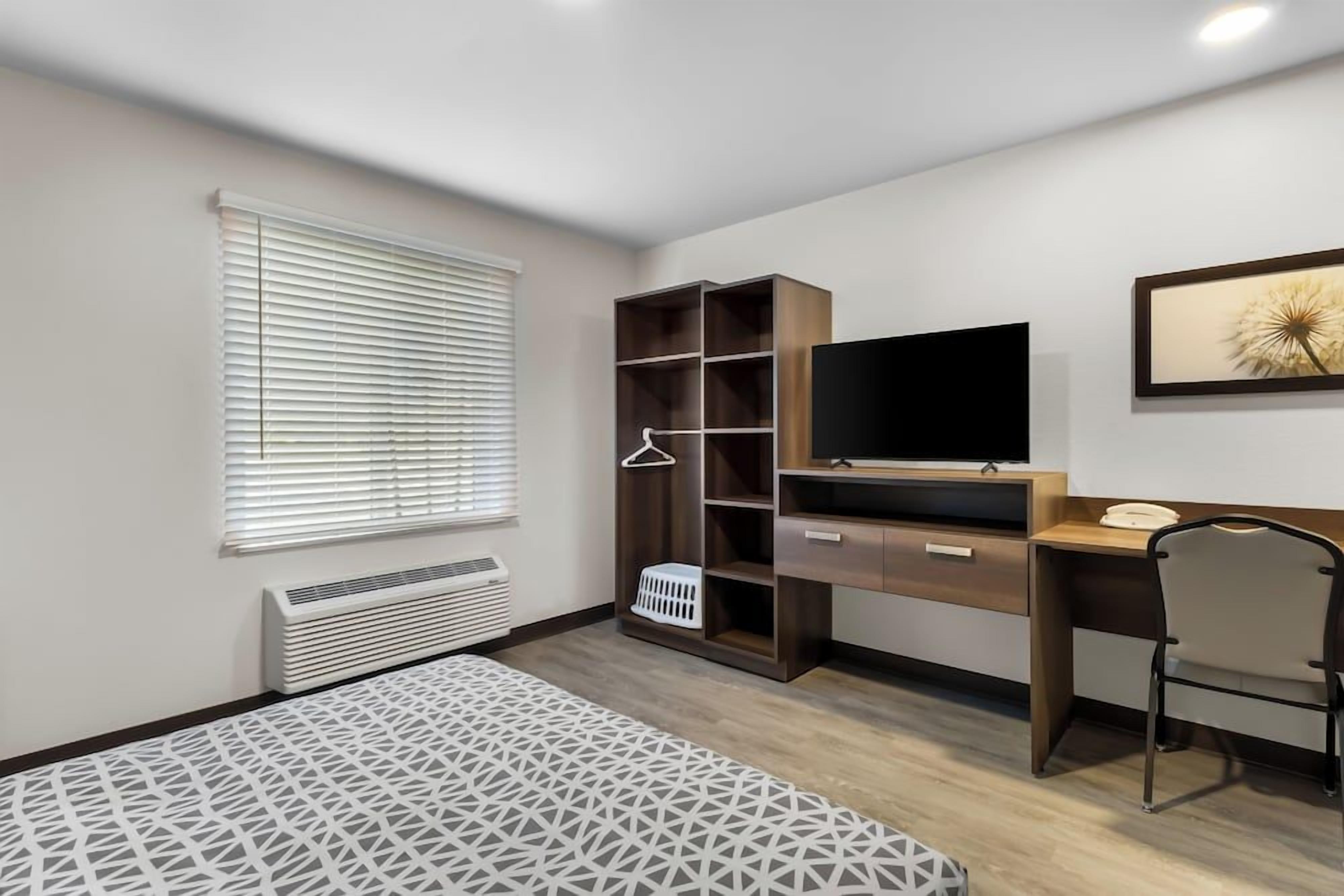 A bedroom with a bed, a desk, a chair, a TV, a wardrobe, and a window with blinds.