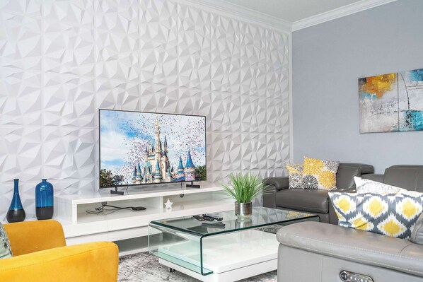 A modern living room with a geometric-patterned wall, a flat-screen TV, a glass coffee table, a sofa, and decorative pillows. - Luxury 6BR Resort Home - Near Disney - Pool, Hot Tub & BBQ! (Kissimmee)