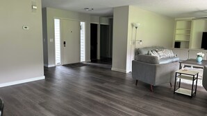 Un salón amplio con un sofá gris, una mesita de centro y una lámpara. Hay una puerta que conduce a otra habitación y un pasillo.
