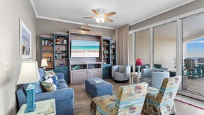 A living room with a large TV, bookshelves, a ceiling fan, and a view through a large window. - 1411 The Tides at Tops'l - Gulf Front, Community Pool, & Kiddie Pool (Miramar Beach)