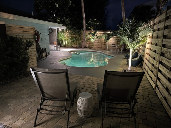 A backyard with a swimming pool, two chairs, a small table, and a wooden fence.