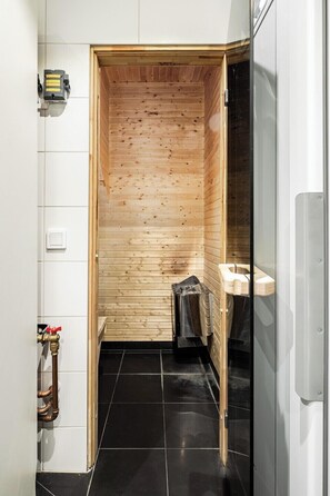 A sauna room with wooden walls, a black tiled floor, and a glass door leading to a bathroom with a toilet and towel rack.