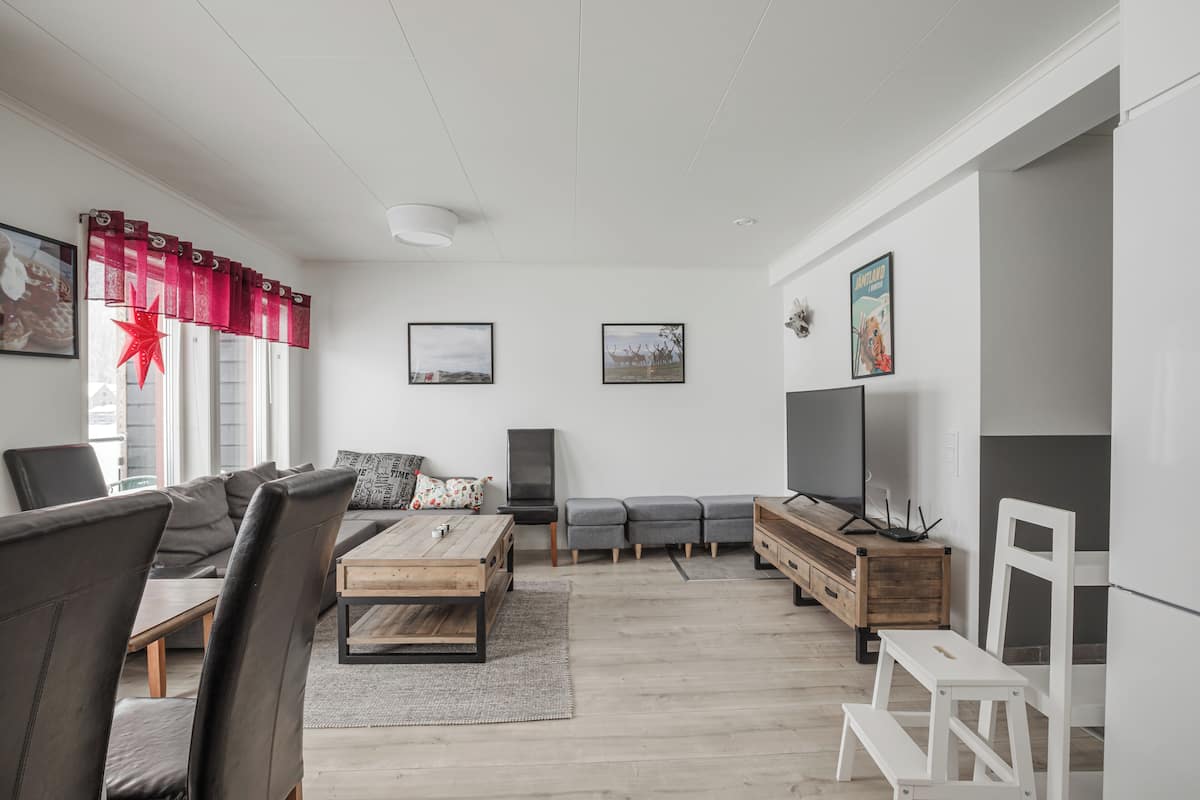 A modern living room with a sofa, coffee table, and TV. There are framed pictures on the wall and a window with red curtains.