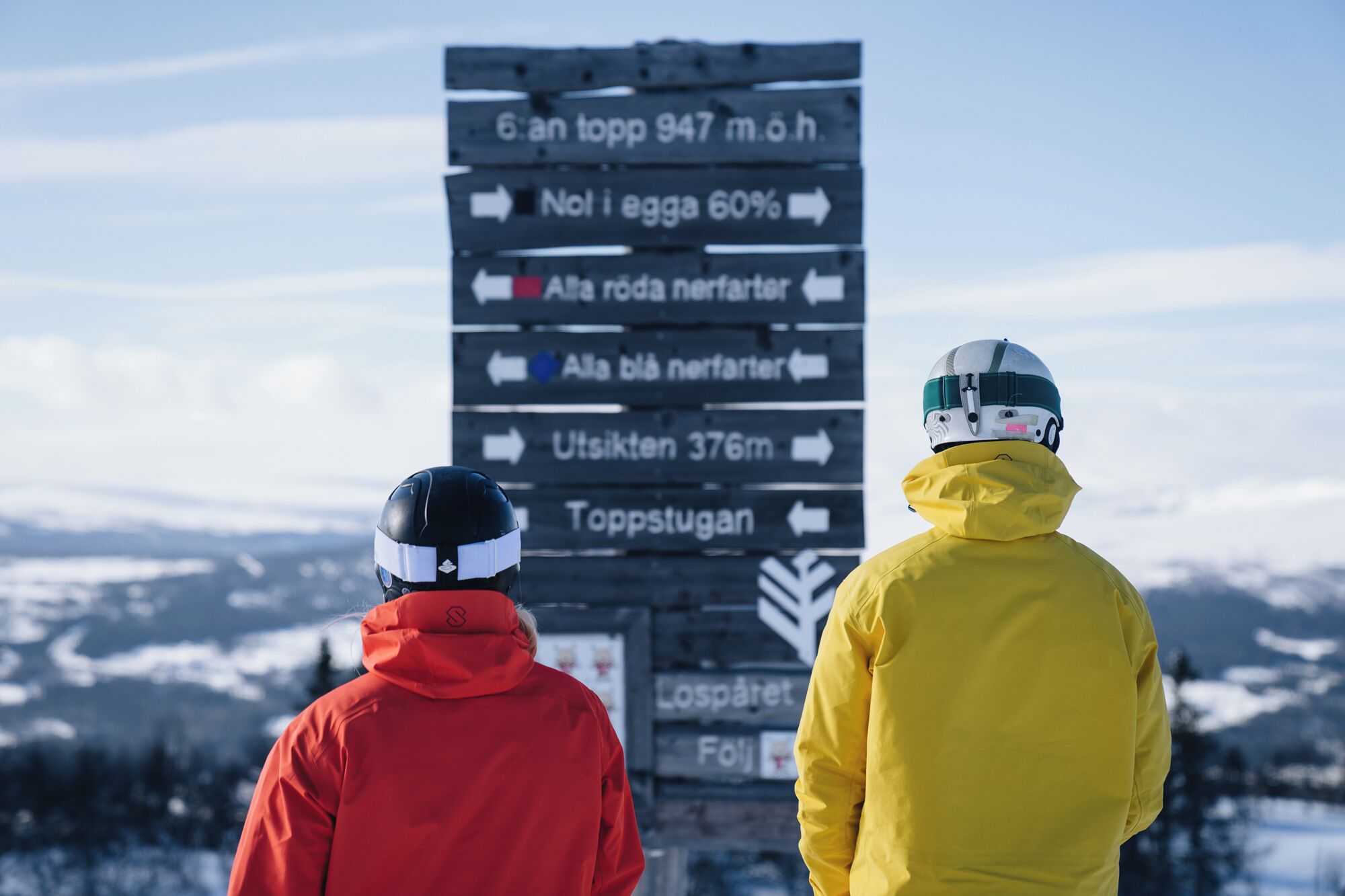 Two people in winter jackets looking at a directional sign with various destinations and distances.