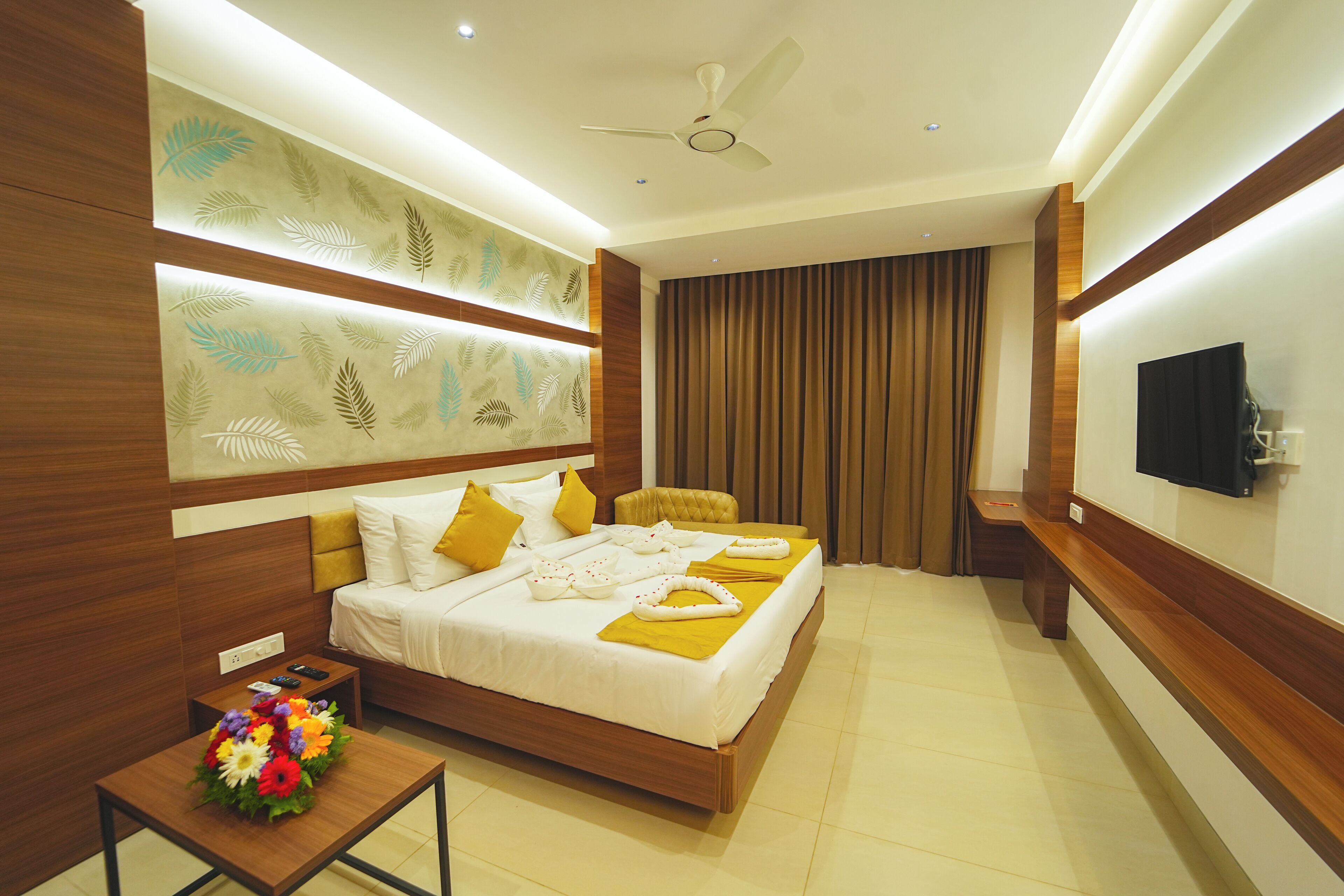 A modern hotel room with a large bed, a flat-screen TV, a ceiling fan, and a decorative wall panel.