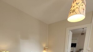 Een net opgemaakt bed met een grijze geweven deken, twee bedlampjes en een hanglamp met bloemenmotief.