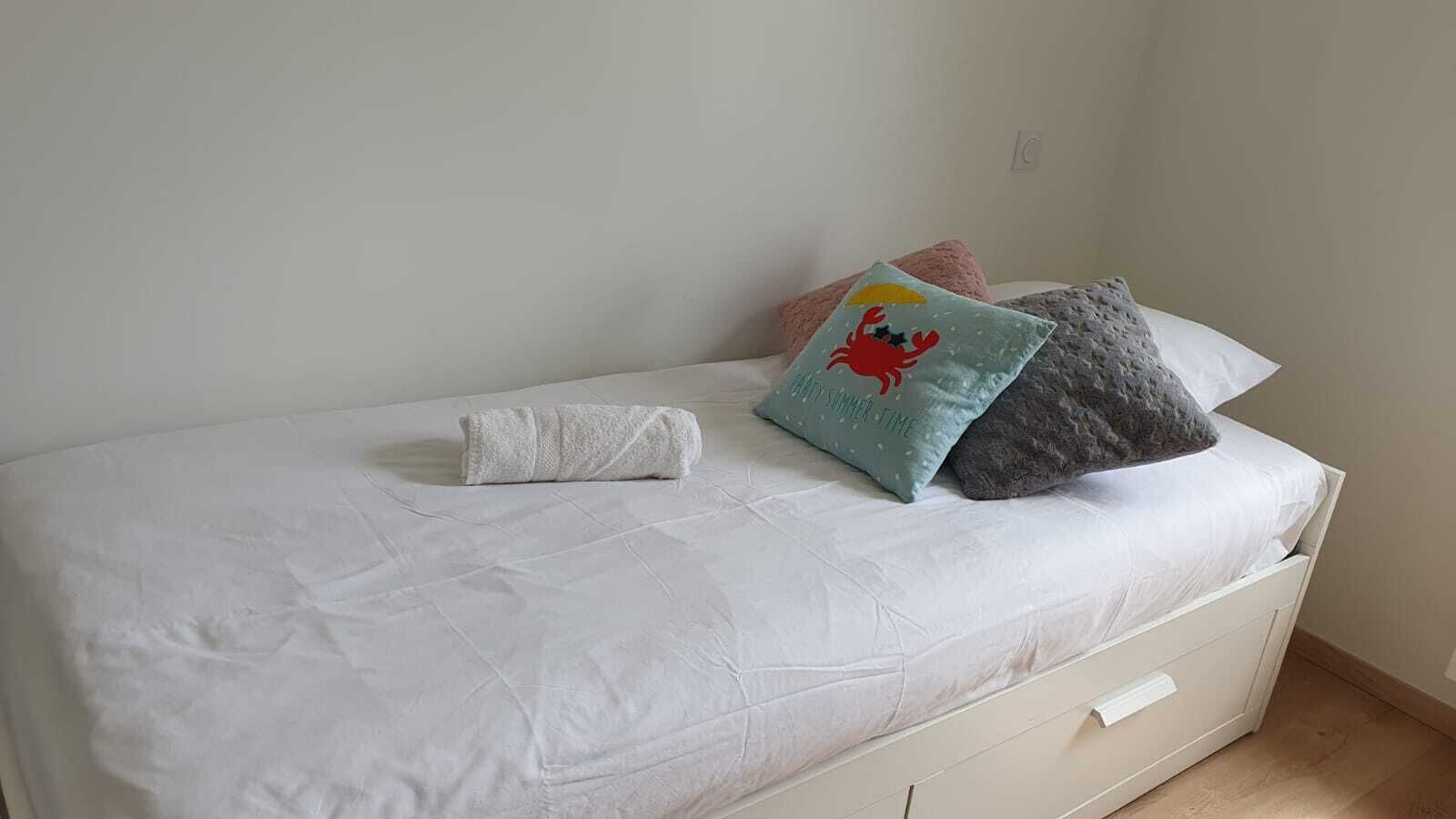 Un lit bien fait, avec une couette blanche, une serviette blanche enroulée et deux coussins décoratifs.