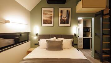 A bedroom with a bed, bedside tables, a headboard, wall-mounted artwork, a ceiling fan, and a kitchenette in the background.