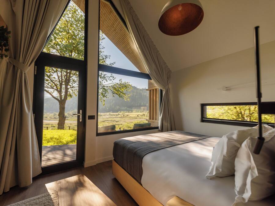 A bedroom with a large window, a bed, and a view of greenery outside.