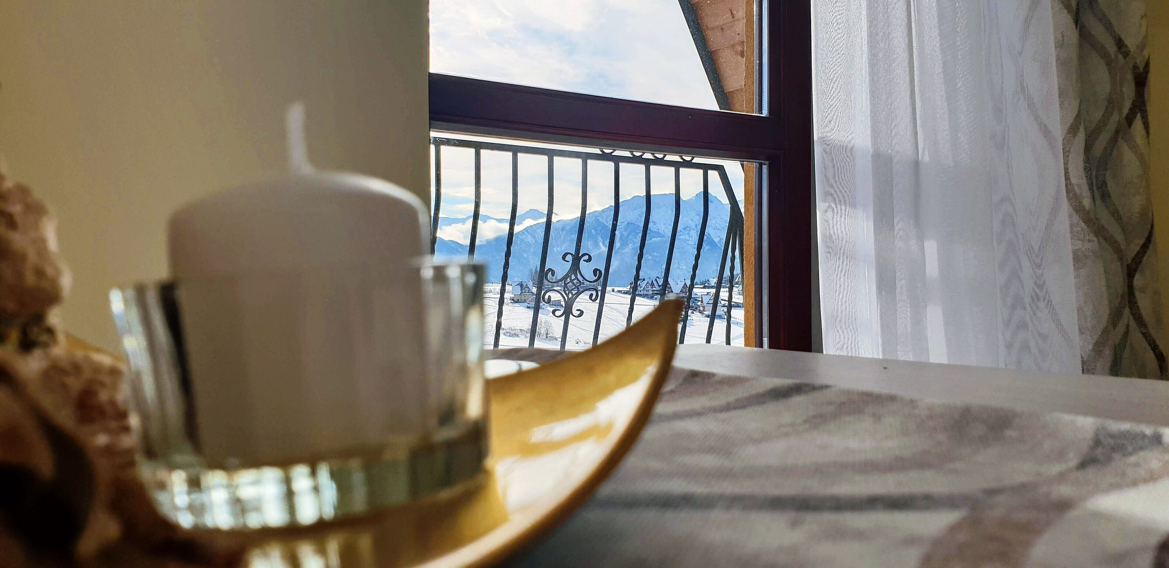 A table with a white cup on a gold saucer, a patterned tablecloth, and a view of mountains through a window with a metal railing.