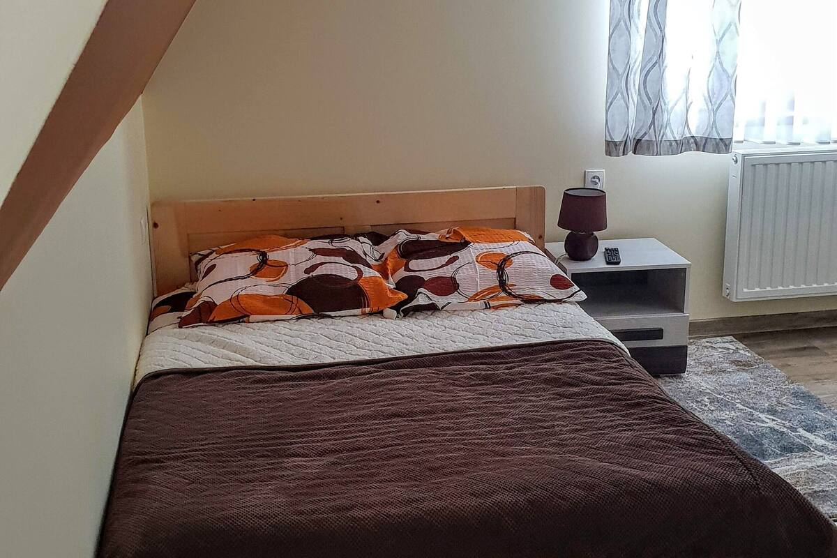 A bedroom with a wooden bed, a bedside table with a lamp, and a window with curtains.