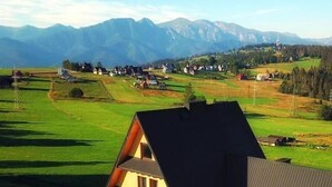 Une maison à plusieurs étages, dotée d’un toit en pente, entourée de champs verdoyants et avec des montagnes en arrière-plan.