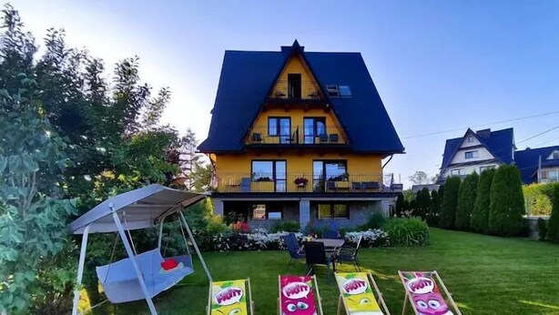 Une maison à deux étages, avec un toit bleu et des murs jaunes, entourée d’une pelouse bien entretenue et d’une balançoire.