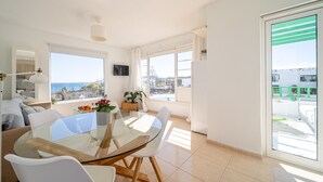 Ein modernes Wohnzimmer mit einem Glastisch, weißen Stühlen und einer Couch. Ein großes Fenster bietet Blick auf das Meer und ein Balkon mit grüner Brüstung.