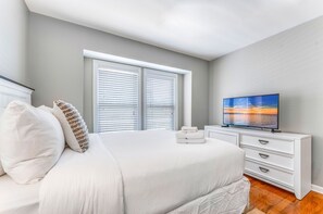 A bedroom with a large bed, white bedding, a TV on a white dresser, and a window with blinds. - Came & Stayed, Community Pool, Tranquility, Beach Gear Credit Included! (St George Island)