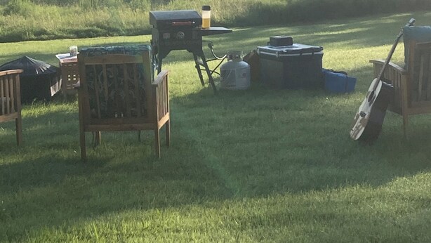 An outdoor area with a barbecue grill, a table with a chair, and a guitar leaning against a wooden structure. - Throw your tent anywhere on property (Westmore)