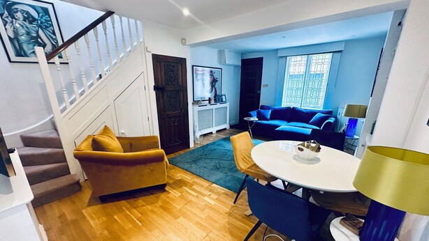 Una sala de estar moderna con una escalera, un sofá azul, un sillón amarillo y un comedor con una mesa y sillas.