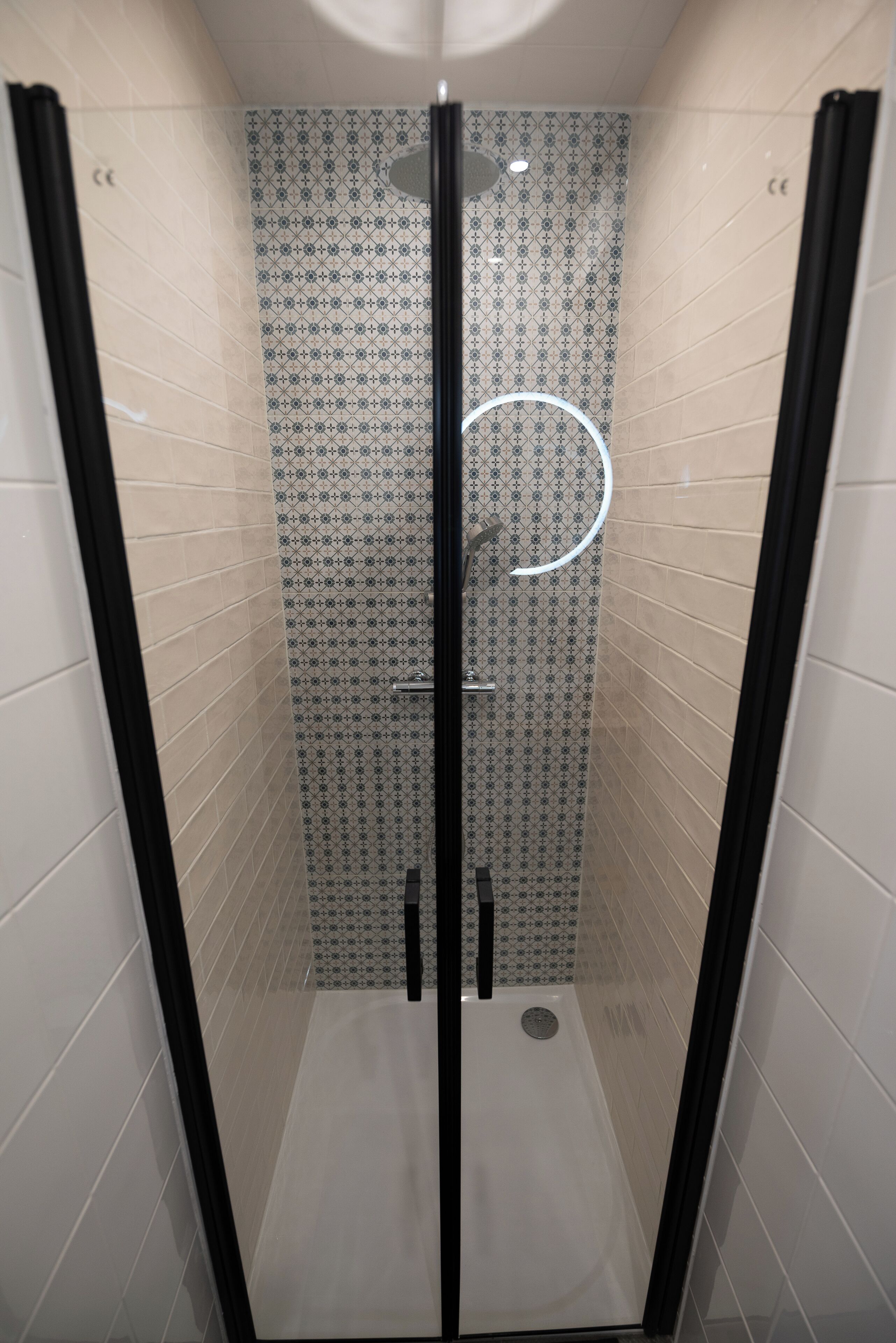 Eine begehbare Dusche mit Glastüren, einer Wand mit gemusterten Fliesen und einer runden Leuchte.