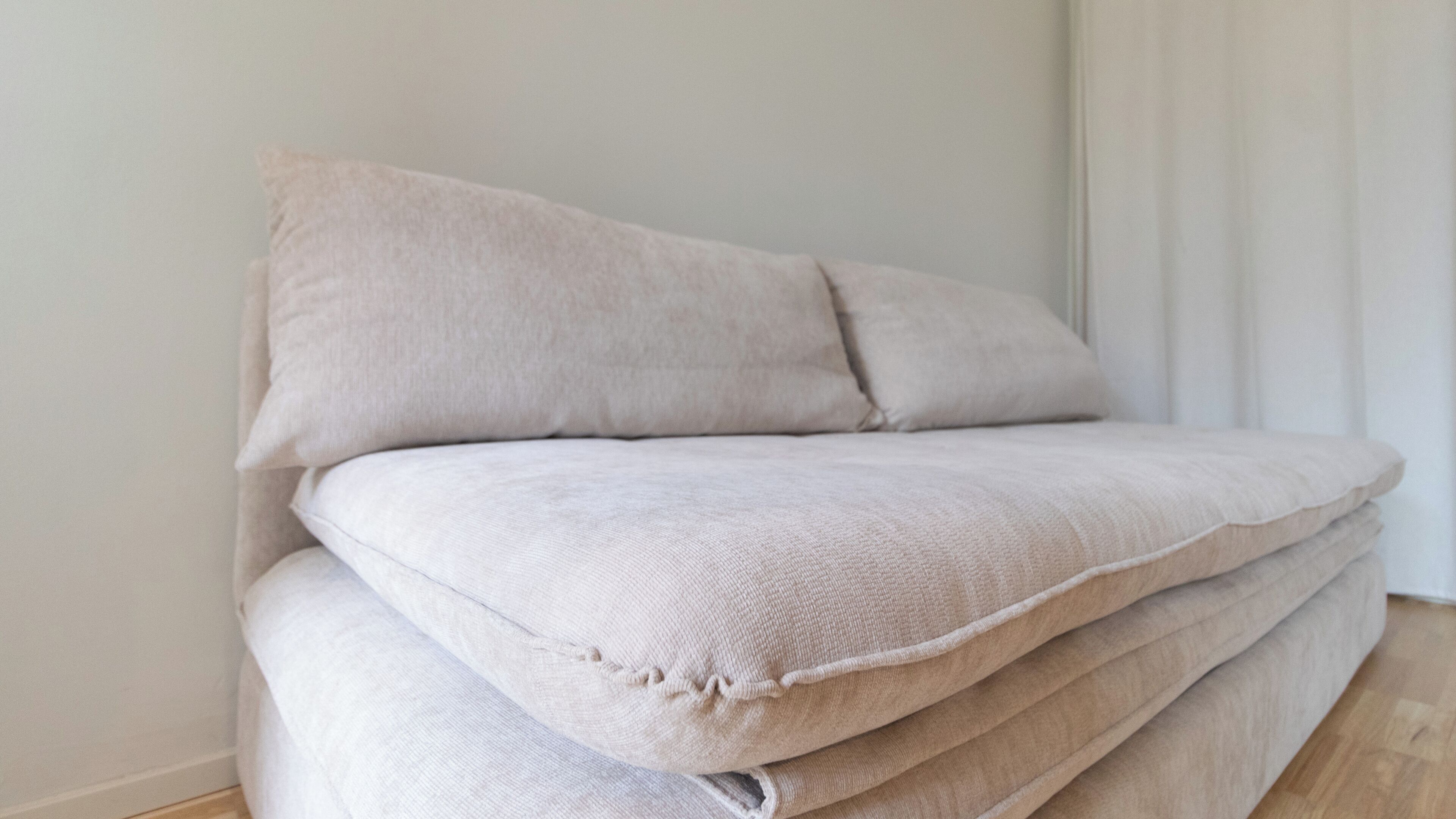 Ein ordentlich gefaltetes Sofa mit heller Stoffbezug und schlichtem Design, platziert vor einer schlichten Wand und einem weißen Vorhang.