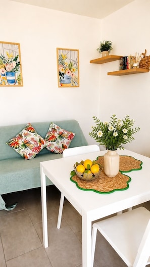 Una sala de estar con un sofá, dos cuadros en la pared, una mesa con un florero y limones, y una pequeña planta en un estante.