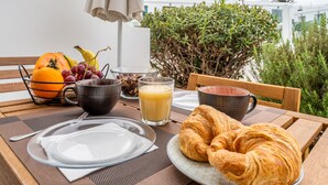 Een ontbijtsetting met croissants, fruit en sap op een tafel met uitzicht op gebouwen en groen.
