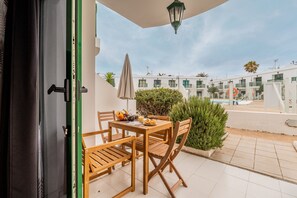 An outdoor patio with a wooden table and chairs, a small table with fruits and a drink, and a view of a pool and buildings in the background. - Sol y Sal de Corralejo (Corralejo)