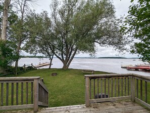 A wooden deck with a bench, a grassy area, and a large tree overlooking a lake with boats. - Waterfront Cottage on Rice lake (Roseneath)