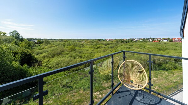 Un balcon agrémenté d’un fauteuil en osier, d’où l’on peut admirer un paysage verdoyant et luxuriant ainsi que des maisons situées au loin.