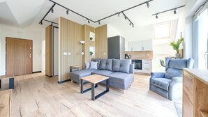 A modern living room with a grey sectional sofa, a wooden coffee table, and a blue armchair. There is a kitchen area with a stove and cabinets in the background. - Apartamenty Sun & Snow Na Różanej (Ustronie Morskie)